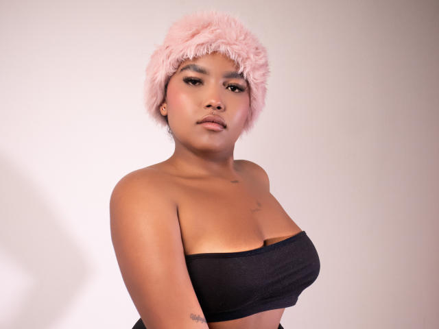 Fotografie von ChrystalFiore, einer attraktiven asiatischen Frau mit kurzen, lockigen rosa Haaren, die ein schwarzes Top ohne Träger trägt. ihr Dekolleté zur Schau stellend, verführerisch in die Kamera blickend mit einem leichten Grinsen.