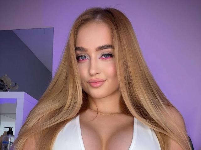 Close-up van ElyssaWetX, een jonge vrouw met lang, steil blond haar, roze oogschaduw en een witte top met een lage halslijn. waarbij haar grote borsten in de schijnwerpers staan. Ze glimlacht met een subtiele, aantrekkelijke blik..