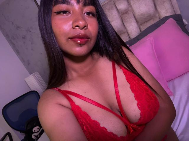 Close-up of NahomyNoir, a curvy woman with long black hair, wearing a red lace bra that accentuates her large breasts. She has a seductive look with glossy lips and pink lighting.