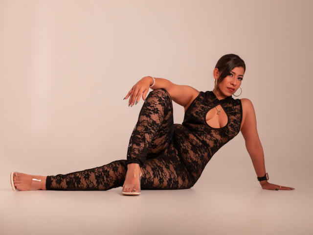 LaurenHills reclines seductively in a black lace bodysuit with a cutout on the chest, legs spread, one arm supporting her while the other rests on her thigh. She wears red high heels.