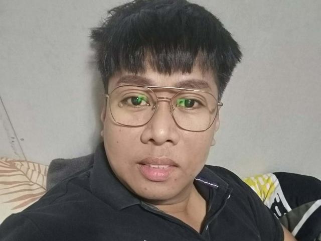 Photograph of PinoYako69 with short black hair, green-tinted glasses, and a black polo shirt. Green eyes, slightly parted pink lips, slight facial stubble. Cock not visible. Casual, direct gaze.