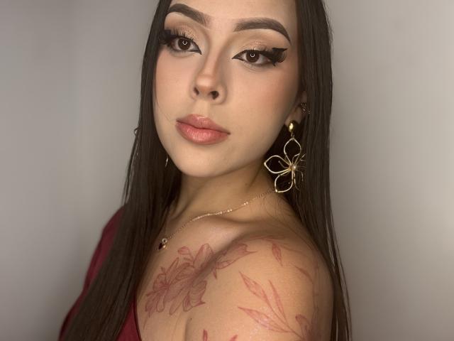 Close-up of AlisonTatcher with long black hair, full lips, and heavy eyeliner. She gazes seductively at the camera, wearing gold earrings, and showing a floral tattoo on her shoulder. Fucking hot.