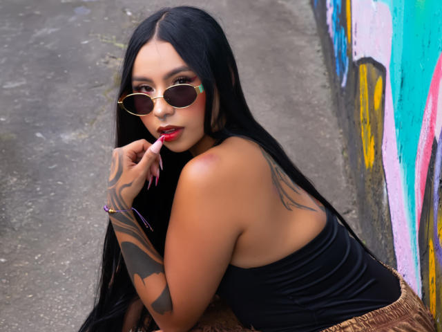 ChloeMoretti, cabelo preto longo, braço tatuado, top preto sem alças, batom vermelho, dedo na boca, óculos de sol redondos, apoiada em uma parede coberta de grafites, sensual, cores vibrantes, ambiente urbano, pose provocativa..