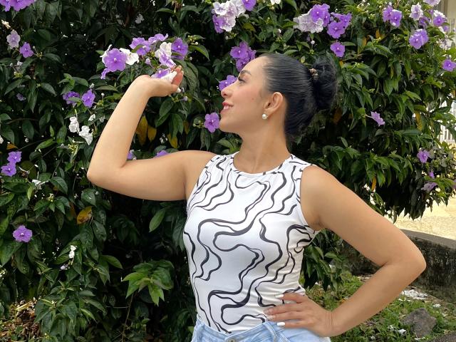 Katty Rivera è in piedi, con una mano appoggiata al fianco, e sta annusando dei fiori viola. Indossa una canotta bianca con un motivo astratto nero. I capelli scuri sono raccolti in uno chignon. Sfondo: fitta vegetazione verde con fiori viola..