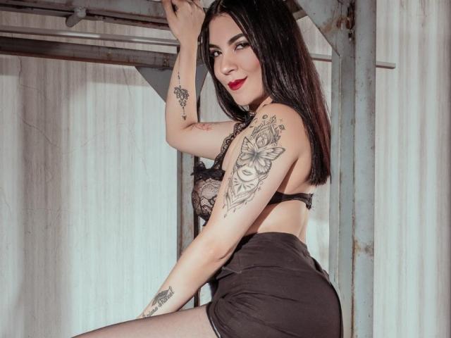 JadeRushh, avec ses cheveux noirs, son rouge à lèvres rouge et ses tatouages, portant un soutien-gorge et un short en dentelle noire, assise sur une structure métallique. Regardant l'appareil photo avec un sourire séduisant. Extrêmement sexy..