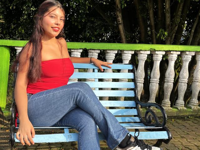 AlyssaGreen zit op een witte houten bank, met gekruiste benen, en draagt een strak rood topje zonder bandjes en blauwe jeans. Ze heeft lang zwart haar, rode lippenstift en zwarte schoenen. Voorgrond: spanning..