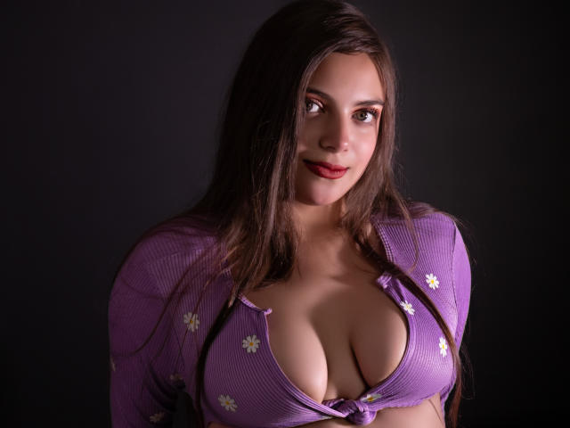 HaleyMolly, com longos cabelos escuros, olha para frente com um leve sorriso. Ela veste uma camisa roxa com decote baixo e flores brancas, que revela seios grandes. Fundo escuro, iluminação suave. Pose sedutora e confiante..