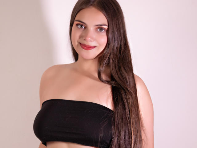 Foto van een jonge vrouw met lang, recht bruin haar, die een zwarte top zonder bandjes draagt en recht in de camera lacht. camera. Ze heeft een lichte huid, rode lippenstift en staat voor een witte achtergrond. Haar lichaam is lichtjes naar rechts gekeerd. Haar borsten zijn van gemiddelde grootte, en er is een subtiele hint van decolleté..