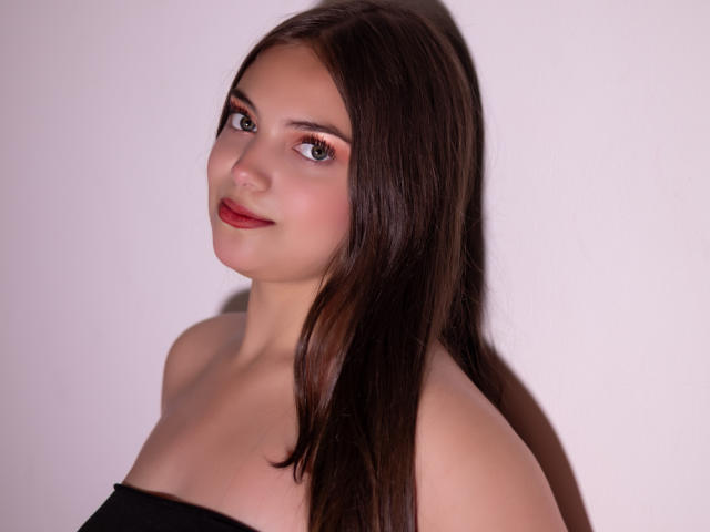 Fotografie einer jungen Frau, HaleyMolly, mit langen, glatten, schwarzen Haaren, heller Haut und rotem Lippenstift. Sie trägt ein schwarzes Top ohne Träger und blickt mit einem leichten Lächeln über ihre Schulter. Ihre Augen sind dunkel und sie hat einen subtilen, verführerischen Blick..