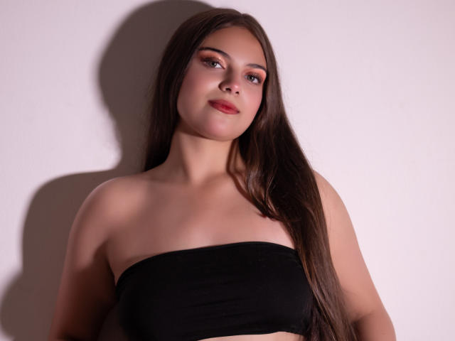 Fotografia di HaleyMolly con lunghi capelli castani lisci, che indossa un top nero senza spalline. Ha la pelle chiara, il rossetto rosso e un'espressione sicura. Una luce intensa proietta un'ombra sul suo lato sinistro..