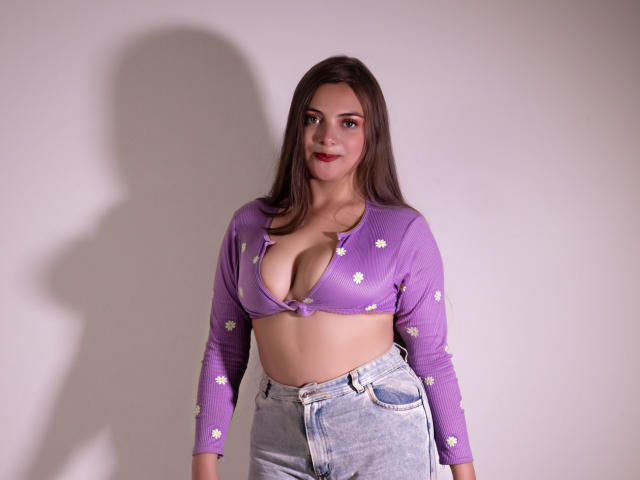 HaleyMolly, übergewichtig mit großen Brüsten, trägt ein lila Crop-Top mit weißen Blumen, das aufgeknöpft ist, um das Dekolleté zu zeigen. und Jeans mit hoher Taille. Schatten an einer weißen Wand. Eine sexy, selbstbewusste Pose..