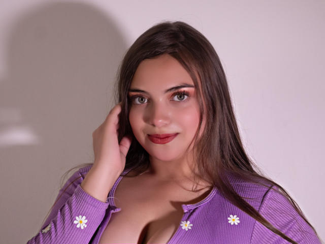 Foto van een mollige vrouw met lang bruin haar, die een paars shirt draagt met witte margrietmotieven, met haar hand voor haar gezicht. rode lippenstift, decolleté zichtbaar, directe blik, zachte belichting.