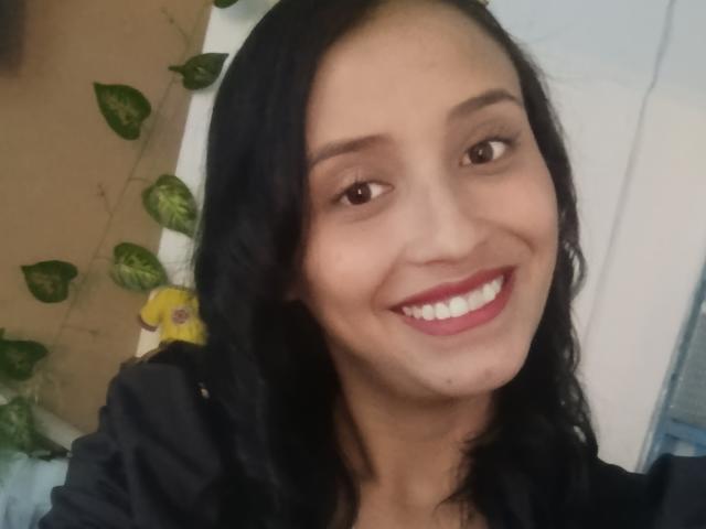 Primer plano de PreatyPepper, con tono de piel medio, cabello negro ondulado y lápiz labial rojo, sonriendo ampliamente. Ella tiene ojos oscuros y una pequeña peca en su mejilla derecha. El fondo incluye hojas verdes y una flor amarilla. Ella lleva una blusa negra. Increíblemente atractiva..