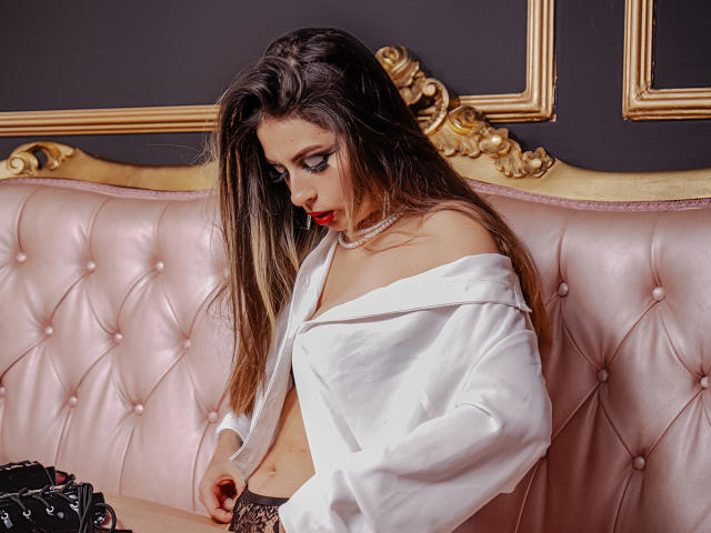 LeilaGoddes sitzt auf einem rosafarbenen, gepolsterten Ledersofa mit goldener Verzierung und trägt ein weißes Top mit einem Schulter frei lassenden Ausschnitt. Sie hat langes, braunes Haar, roten Lippenstift und eine Perlenkette und blickt nach unten. Ihre Pose ist verführerisch, mit einem Hauch von Anspannung..