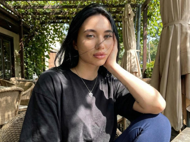 Photograph of AlexaStrei, an Asian woman with long black hair, wearing a black t-shirt and blue jeans, sitting outdoors, hand on cheek, sunlit, shadows on face, relaxed, slightly seductive.