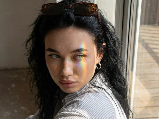Photograph of AlexaStrei with long black hair, wearing a white shirt, standing near a window. Sunlight creates colorful reflections on her face. Eyeglasses on head, hoop earrings. Sexy, intense gaze.