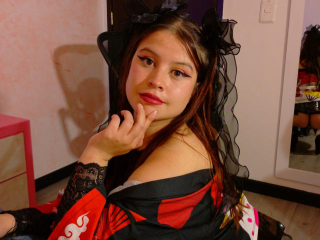 MelaniBecker with long brown hair, black lace headpiece, and red-black kimono, sitting, hand on chin, looking directly at camera, slight smile, shadow on wall, casual indoor setting.