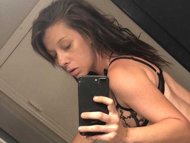 Autoritratto ravvicinato di LenaRae, con lunghi capelli castani, che indossa un reggiseno nero in pizzo, tiene uno smartphone nero e guarda verso... fotografia di una donna con gli occhi socchiusi, che mostra leggermente il seno, riflessa in uno specchio..