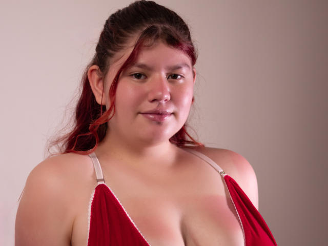 Fotografie von Karla Harper, einer übergewichtigen Frau mit heller Haut und roten Haaren, die ein tief ausgeschnittenes rotes Kleid trägt, das dekolleté. Sie hat ein dezentes, selbstbewusstes Lächeln. Weiches Licht betont ihre Rundungen..