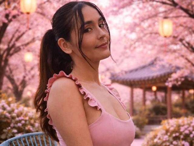 Fotografia di MindyLove, una giovane donna con lunghi capelli neri raccolti in una coda di cavallo, che indossa una canotta rosa con volant, in piedi. Un giardino di alberi di ciliegio in fiore. Sorriso dolce, lanterne sfocate sullo sfondo.