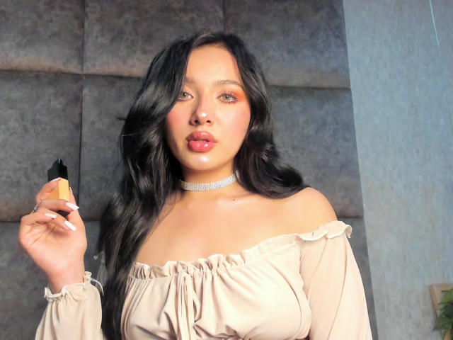 LillyTaylor, an attractive Asian woman with long black hair, holds a black object in her right hand. She wears a white off-shoulder top and a choker. Her red lips are slightly parted.