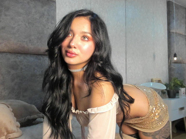 LillyTaylor with long black hair, leaning forward on a bed, wearing a white off-shoulder top and gold sequined skirt, highlighting her curvy figure and visible tattoo on her hip. Sunlight highlights her face.