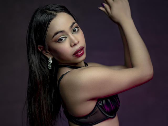 CarlaLordes, with dark straight hair, raises her right arm, wearing a black bra, deep red lipstick, and dangling earrings. Sultry gaze, glossy skin, intense sexual tension, dark background.