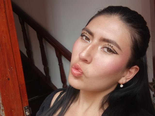 Photograph of EmmaRouxx with glossy pink lips puckered in a kiss, brown eyes with long lashes, pink blush on cheeks, dark hair pulled back, wearing black top, diamond earring, wooden railing background.