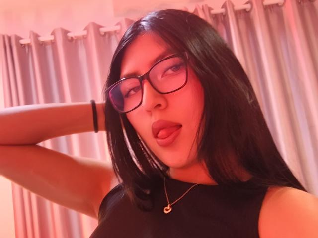 Close-up selfie van EmmaRouxx, een aantrekkelijke vrouw met lang zwart haar, die zwarte bril en een zwarte top draagt. Ze steekt haar tong uit en heft een arm. Rood licht, gordijnen op de achtergrond..