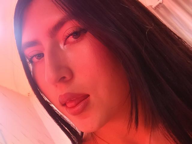 Close-up van EmmaRouxx met rood licht, donker, recht haar, volle lippen die lichtjes open zijn, bruine ogen die recht in de camera kijken. de camera, een zachte gloed op haar gezicht, een lichte schaduw op haar rechterwang..