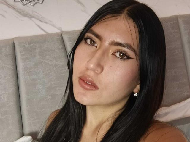 Photo en gros plan d'EmmaRouxx, une femme séduisante avec de longs cheveux noirs, des yeux marron et des lèvres pulpeuses, qui regarde directement l'objectif. caméra. Elle a une expression séduisante, et elle porte un haut marron. Arrière-plan gris avec une tête de lit..