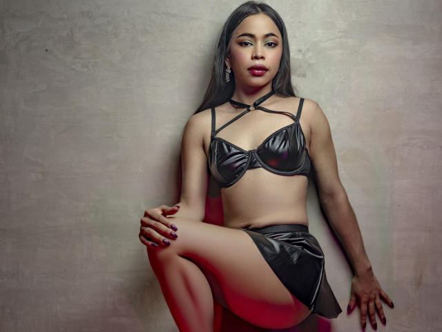 CarlaLordes leans against a gray wall, wearing black leather lingerie with strappy details. She has long black hair, red nails, and a serious expression. One leg is raised. Sexual tension is palpable.