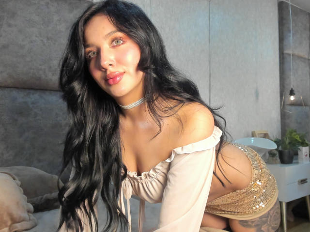 LillyTaylor leans forward on a bed, black hair cascading, white off-shoulder top, gold sequin skirt, choker necklace. Sunlight highlights her face, curvy figure, and thick thighs. Sexual tension evident.