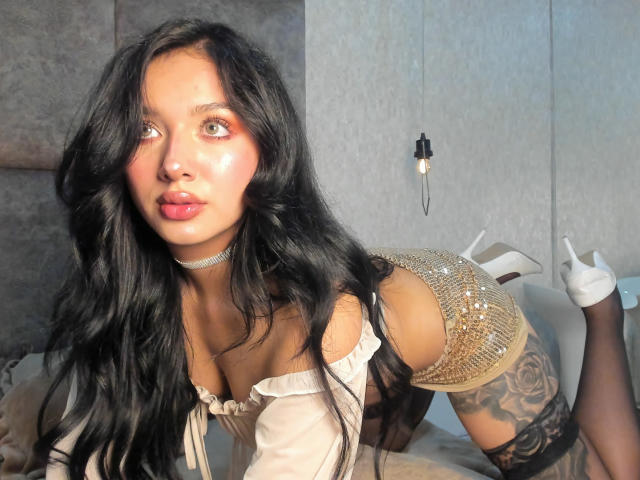 LillyTaylor with long black hair, red lipstick, and a sparkling choker, on all fours in a white dress, showcasing a tattooed thigh and wearing white high heels. Fucking sexy pose.