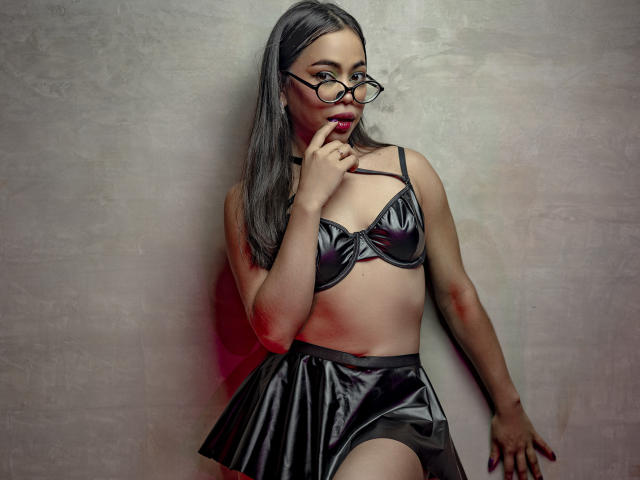 CarlaLordes stands against a textured gray wall, wearing black lingerie with a shiny bra and pleated skirt. She has long black hair, glasses, and a finger in her mouth. Fucking sexy.