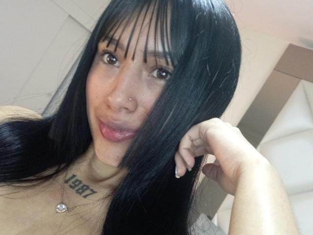 Fotografía de primer plano de SkarletAry, una joven con cabello largo y negro, flequillo y ojos marrones, que lleva un piercing en la nariz y una collar con un dije en forma de corazón. El tatuaje en el cuello dice "LILY". Increíblemente sexy, mirando a la cámara con una ligera sonrisa..