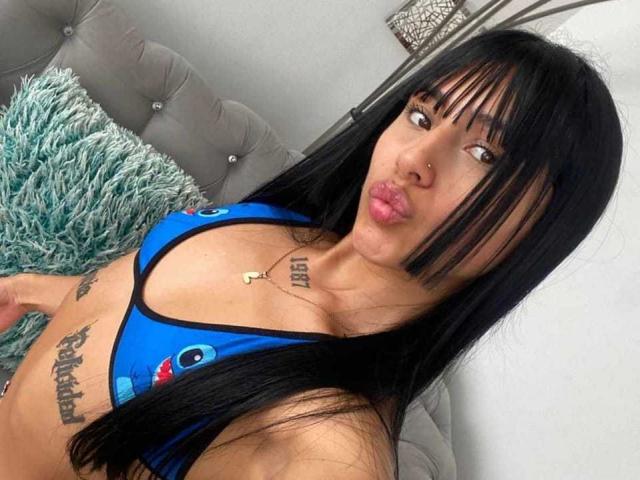 Fotografia di SkarletAry, una donna asiatica con lunghi capelli neri, che indossa un top da bikini blu con un motivo Pac-Man, sdraiata su un divano grigio con un cuscino blu peloso. I suoi tatuaggi sono visibili sul petto e sul fianco. Ha un piercing al naso e sta facendo la smorfia. I suoi occhi guardano a destra
.