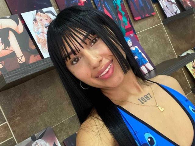 Gros plan de SkarletAry, une femme souriante avec de longs cheveux noirs, une frange et des yeux marrons, portant un haut bleu à encolure américaine, montrant un tatouage "BBB" sur sa poitrine, dans une pièce décorée avec des œuvres d'art mural colorées..