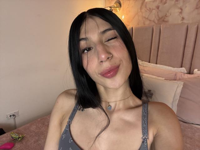 Fotografia de Sophie Beaumont com cabelo preto, piscando, vestindo uma regata cinza, mostrando o decote, usando um colar e um piercing no nariz. Sorrindo, em um quarto com uma cabeceira rosa, uma luminária e iluminação suave. Muito fofo.