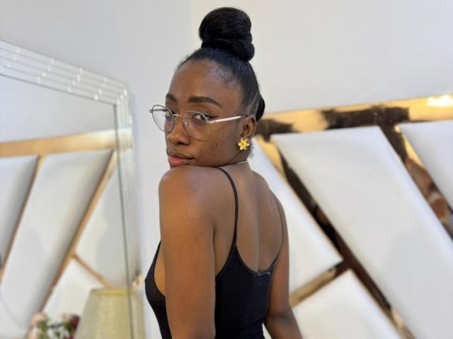 Photographie de SelenaD, avec une peau foncée, des cheveux noirs attachés en chignon, portant des lunettes et un haut noir à bretelles fines, regardant en arrière. par-dessus son épaule, poutres en bois en arrière-plan, pose sexy.