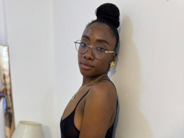 Fotografía de SelenaD, una mujer negra atractiva de piel oscura, que lleva gafas y una camiseta negra sin mangas, con el pelo recogido. moño alto, de pie frente a una pared blanca, mirando seductoramente a la cámara.