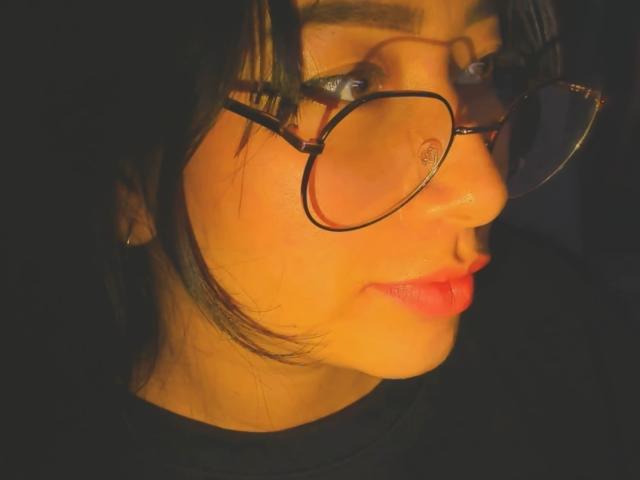 Close-up of Genesiss, side profile, with glowing yellow light on face, black hair, round glasses, red lips, black shirt, intense gaze, shadows, high contrast, sexy, sultry vibe, warm light.