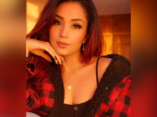 Fotografia di KattyWattson con capelli castano di lunghezza media, che indossa una camicia a quadri rossa e nera, una maglia nera e un accessorio dorato. collana.
lei appoggia il mento sulla mano, con un'espressione sensuale. Illuminazione calda e arancione..