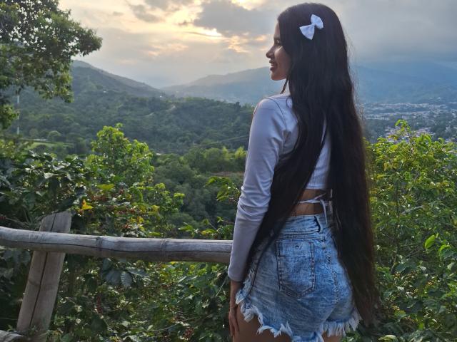ScarlettRoss69 está em um balcão de madeira, de costas, com cabelos longos e pretos, uma blusa branca e shorts jeans desgastados. O sol se põe sobre montanhas exuberantes, projetando um brilho suave. Uma vista incrivelmente linda..