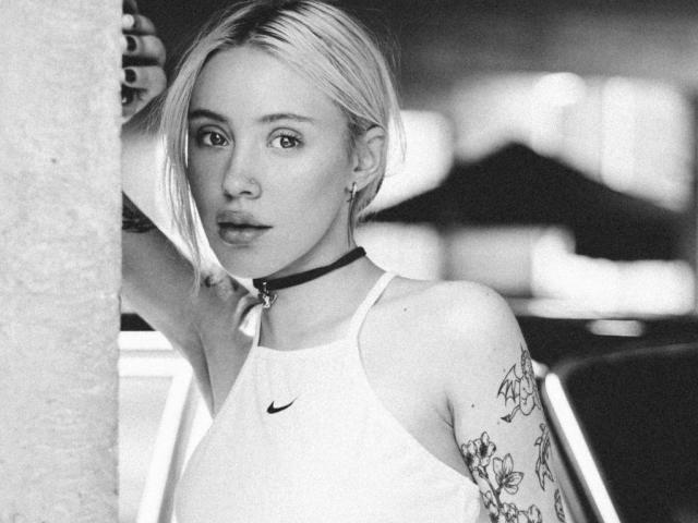 Zwart-wit foto van WandaMchardy, met kort blond haar, die een choker en een Nike tanktop draagt, met een tatoeage op haar linkerarm. Intense blik, staand naast een pilaar, heldere achtergrond. Heel aantrekkelijk..