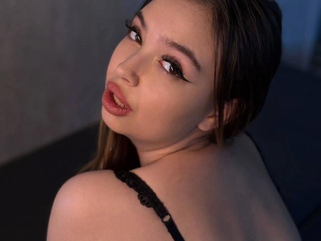 Primo piano di JullyMood: una donna con la pelle chiara, capelli lunghi castani, eyeliner nero e rossetto rosso, che guarda oltre la telecamera. con un'espressione seducente. Indossando una camicetta di pizzo nero, illuminazione soffusa..