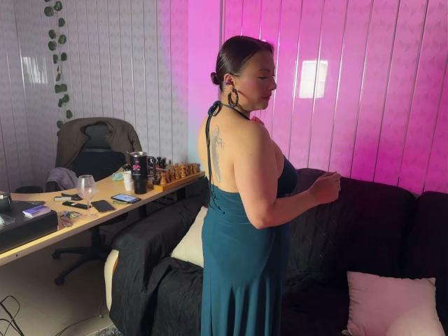 SusanaHotSex, de perfil, vestida con un vestido negro de tirantes, mostrando un tatuaje en la parte superior de la espalda. Habitación iluminada con luz rosa, con un escritorio, un tablero de ajedrez, una silla y un mando de videojuegos. Tensión sexual.