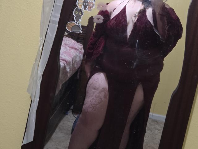 Fotografia de JackVelis em um espelho, vestindo um vestido vermelho escuro com uma fenda alta na coxa, revelando renda estampada. meias. A luz refletida cria um efeito brilhante. Pose incrivelmente atraente..