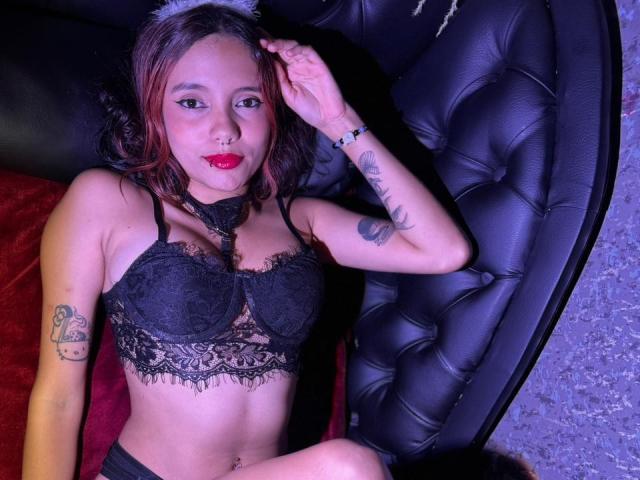 AmarandSofty, avec ses cheveux bruns et son rouge à lèvres rouge, portant un soutien-gorge en dentelle noire, assise sur un canapé en cuir noir capitonné. Tattoos visibles sur le bras droit, main sur la tête, lèvres rouges, expression sexy..