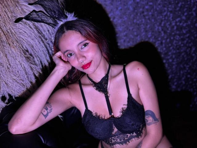 AmarandSofty, vêtue d'un soutien-gorge en dentelle noire et de cottails, se penche sur une surface poilue, une main sur la tête. Peau pâle, rouge à lèvres rouge, tatouages sur les bras. Éclairage violet-bleu, séduisant..